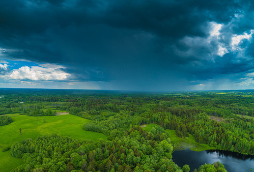 Aerial view of a lush green landscape featuring dense forests open green fields a small dark lake and a dramatic dark stormy sky with rain clouds in Latvia. Aerial view of a lush green landscape featuring dense forests open green fields a small dark lake and a dramatic dark stormy sky with rain clouds in Latvia.