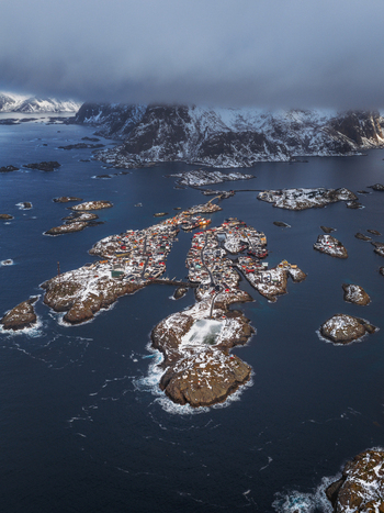 Aerial view of Henningsv&aelig;r s snow dusted buildings nestled among rocky islands contrasted against the deep blue sea and the distant cloud veiled mountains Henningsv&aelig;r Nordland Norway.