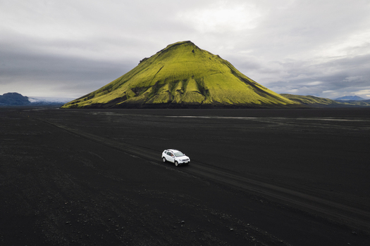 Aerial view of a solitary white vehicle navigates the stark black volcanic landscape towards the vibrant green mossy Mælifell volcano against a moody sky Maelifell Iceland. Aerial view of a solitary white vehicle navigates the stark black volcanic landscape towards the vibrant green mossy Mælifell volcano against a moody sky Maelifell Iceland.