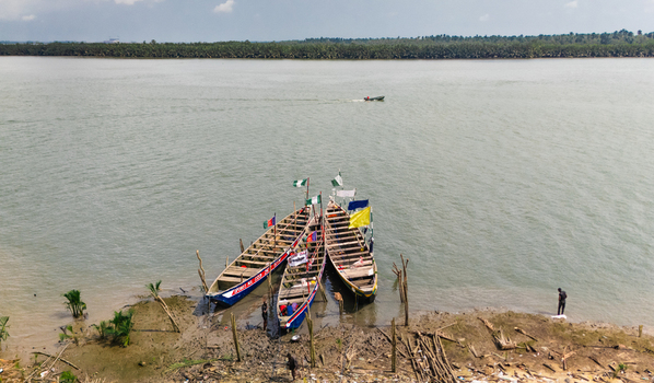 Aerial view of traditional wooden boats moored at the muddy riverbank with flags a small motorboat in the distance and people standing on the shore in Minima Rivers Nigeria.