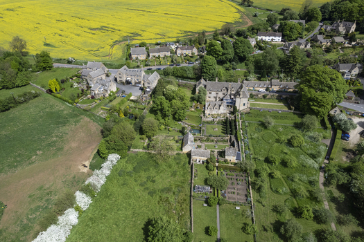 Aerial view of Snowshill Manor with its historic stone buildings formal gardens and lush green fields set against vibrant yellow rapeseed crops in Snowshill England United Kingdom.
