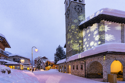 View of the stone church tower with snowflake light projections and snow covered rooftops at dusk in Madesimo Lombardy Italy. View of the stone church tower with snowflake light projections and snow covered rooftops at dusk in Madesimo Lombardy Italy.