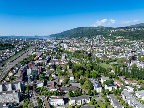 Aerial view of the urban landscape of Biel Bienne featuring residential areas lush green parks and railway tracks leading towards Lake Biel under a clear sky Biel Bienne Canton of Bern Switzerland.