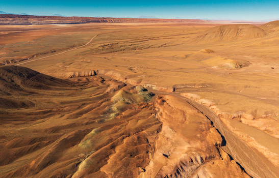 Aerial view of the arid desert landscape with rolling hills deep canyons and a winding dirt road stretching across the vast terrain in San Pedro de Atacama Antofagasta Chile. Aerial view of the arid desert landscape with rolling hills deep canyons and a winding dirt road stretching across the vast terrain in San Pedro de Atacama Antofagasta Chile.