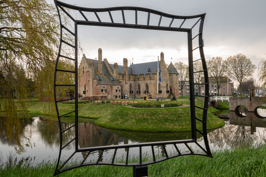 View of Radboud Castle framed by a decorative metal sculpture overlooking a calm moat with green grassy banks and willow trees in Medemblik Noord Holland Netherlands. View of Radboud Castle framed by a decorative metal sculpture overlooking a calm moat with green grassy banks and willow trees in Medemblik Noord Holland Netherlands.