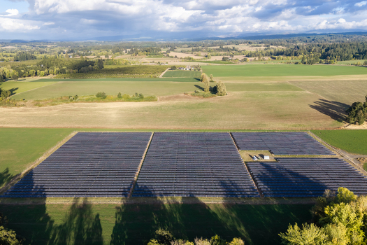 Aerial view of a large solar farm installation set within a rural landscape of green fields and trees under a cloudy blue sky in Woodburn Oregon United States. Aerial view of a large solar farm installation set within a rural landscape of green fields and trees under a cloudy blue sky in Woodburn Oregon United States.