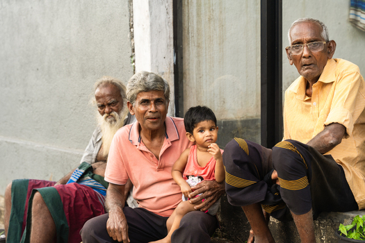 Sri Lanka 15 January 2026 View of a group of elderly Sri Lankan men and a young child sitting together outdoors against a grey wall.