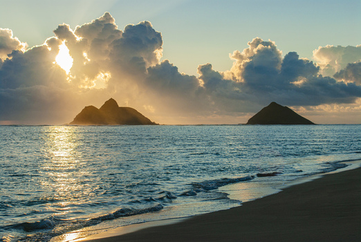 View of the Mokulua Islands at sunrise from Kailua Beach with golden light reflecting on the ocean waves and dramatic clouds in the sky in Kailua Hawaii United States.