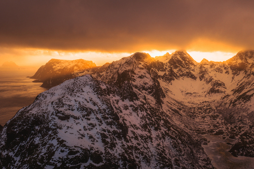 Aerial view of snow capped mountain peaks piercing through a golden sunrise painting the landscape in hues of orange and white Leknes Nordland Norway.