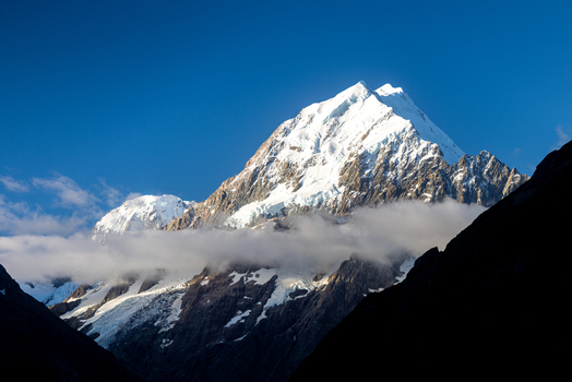 View of a majestic snow capped mountain peak piercing the clear blue sky partially veiled by ethereal clouds Aoraki Mount Cook Canterbury Region New Zealand. View of a majestic snow capped mountain peak piercing the clear blue sky partially veiled by ethereal clouds Aoraki Mount Cook Canterbury Region New Zealand.
