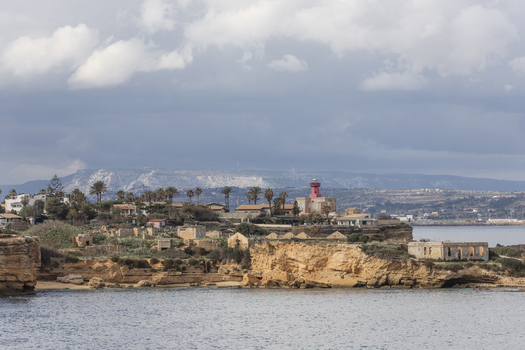View of the red Capo Murro di Porco Lighthouse on the rocky cliffs with abandoned stone structures and palm trees Syracuse Sicily Italy.
