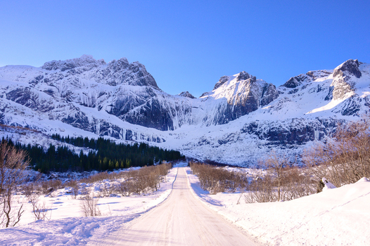View of a snow covered road leading towards majestic snow capped mountains under a clear blue sky in Leknes Lofoten Norway.