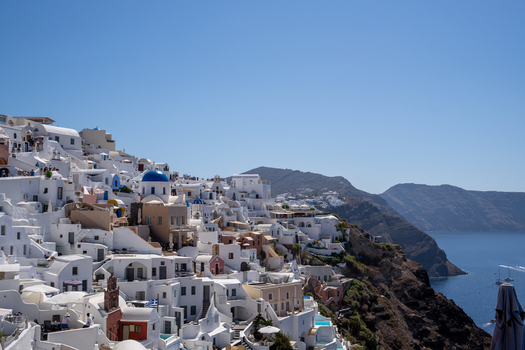 View of the white houses and blue domed churches of Fira on the volcanic cliffs overlooking the Aegean Sea under a clear blue sky in Fira Greece. View of the white houses and blue domed churches of Fira on the volcanic cliffs overlooking the Aegean Sea under a clear blue sky in Fira Greece.