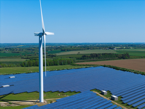 Aerial view of Chelveston Wind Farm featuring a large wind turbine and extensive solar panel arrays set against green fields and a clear blue sky in Wellingborough England United Kingdom. Aerial view of Chelveston Wind Farm featuring a large wind turbine and extensive solar panel arrays set against green fields and a clear blue sky in Wellingborough England United Kingdom.