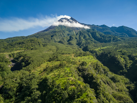 Aerial view of Mount Merapi volcano with smoke rising from the crater and lush green slopes under a clear blue sky in Kecamatan Cangkringan Daerah Istimewa Yogyakarta Indonesia.