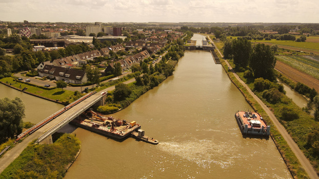 Aerial view of a barge navigating the canal under a bridge with residential houses and green fields lining the banks in Saint Omer Hauts de France France. Aerial view of a barge navigating the canal under a bridge with residential houses and green fields lining the banks in Saint Omer Hauts de France France.
