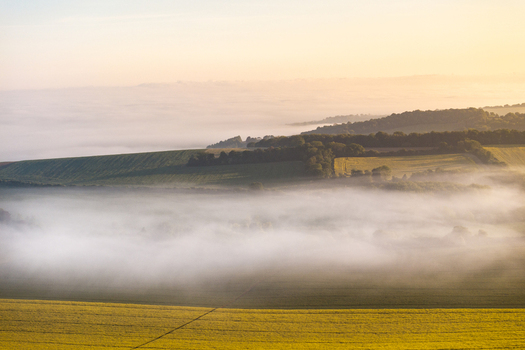 Aerial view of misty green fields and rolling hills covered in dense fog under soft golden morning light in Nuffield Oxfordshire United Kingdom. Aerial view of misty green fields and rolling hills covered in dense fog under soft golden morning light in Nuffield Oxfordshire United Kingdom.