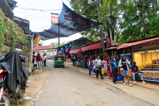 Ratnapura Sri Lanka 22 February 2026 View of a busy street near Adam s Peak with people walking past local shops and a green tuk tuk under a Buddhist flag.