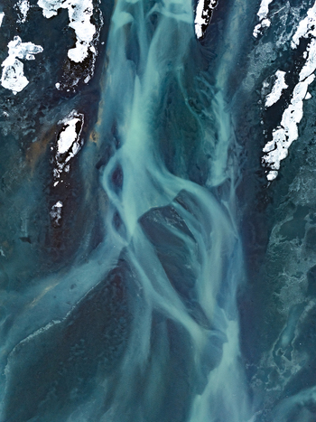 Aerial view of a braided glacial river with turquoise sediment rich water flowing through dark volcanic terrain and patches of snow in Iceland. Aerial view of a braided glacial river with turquoise sediment rich water flowing through dark volcanic terrain and patches of snow in Iceland.