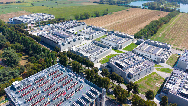 Aerial view of a modern data center campus featuring multiple server buildings with rooftop cooling units surrounded by green trees and agricultural fields in Milan Lombardy Italy. Aerial view of a modern data center campus featuring multiple server buildings with rooftop cooling units surrounded by green trees and agricultural fields in Milan Lombardy Italy.