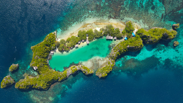 Aerial view of a tropical island with a turquoise lagoon lush green vegetation and small wooden huts along the shore surrounded by deep blue ocean water in Indonesia. Aerial view of a tropical island with a turquoise lagoon lush green vegetation and small wooden huts along the shore surrounded by deep blue ocean water in Indonesia.