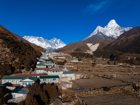 Aerial view of a Himalayan village with stone houses and colorful roofs set against the snow capped Ama Dablam peak surrounded by terraced fields under a clear blue sky in Nepal.