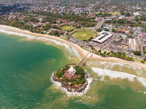 Aerial view of Parevi Duwa Temple on a small island connected by a bridge to the sandy beach and urban coastline in Matara Southern Province Sri Lanka. Aerial view of Parevi Duwa Temple on a small island connected by a bridge to the sandy beach and urban coastline in Matara Southern Province Sri Lanka.