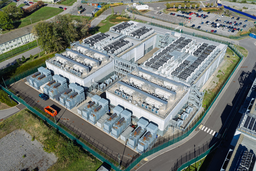 Aerial view of a large data center facility featuring rows of cooling units industrial server buildings security fencing and adjacent parking areas in the United Kingdom.