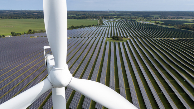 Aerial view of a wind turbine blade in the foreground overlooking a vast solar farm with rows of photovoltaic panels in a rural landscape in Poland.