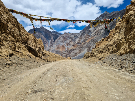 View of the rugged Penzi La road featuring colorful prayer flags strung across the path against a backdrop of towering mountain peaks under a cloudy sky in Zanskar Ladakh India.