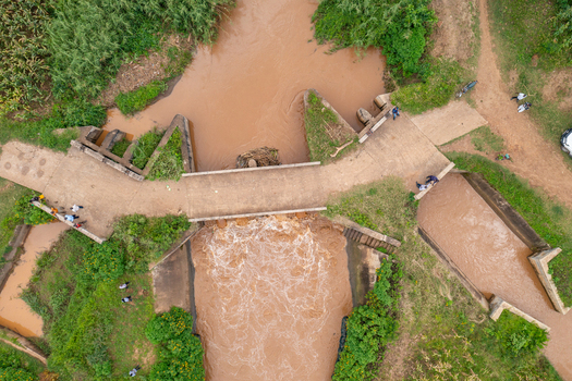 Aerial view of a concrete bridge spanning a muddy river in a rural landscape with lush vegetation and people walking nearby in Butaleja Eastern Region Uganda. Aerial view of a concrete bridge spanning a muddy river in a rural landscape with lush vegetation and people walking nearby in Butaleja Eastern Region Uganda.