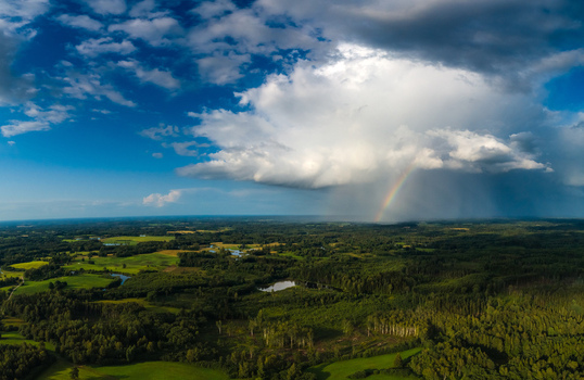 Aerial view of a lush green landscape featuring a dramatic storm cloud and a vibrant rainbow stretching across the horizon in Latvia. Aerial view of a lush green landscape featuring a dramatic storm cloud and a vibrant rainbow stretching across the horizon in Latvia.