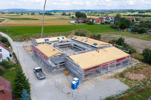 Igensdorf Germany 11 May 2024 Aerial view of a large building under construction with scaffolding a crane arm and workers on the roof surrounded by rural fields and houses. Igensdorf Germany 11 May 2024 Aerial view of a large building under construction with scaffolding a crane arm and workers on the roof surrounded by rural fields and houses.