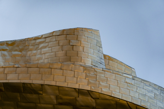 View of the curved titanium panels of the Guggenheim Museum Bilbao reflecting golden light against a clear blue sky in Bilbao Euskadi Spain. View of the curved titanium panels of the Guggenheim Museum Bilbao reflecting golden light against a clear blue sky in Bilbao Euskadi Spain.