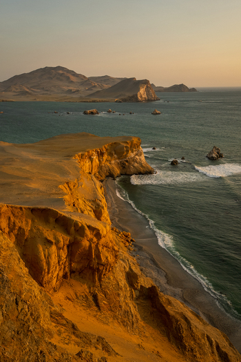 View of the rugged cliffs and golden coastline of Paracas National Reserve at sunset with waves crashing against the shore in Paracas Ica Peru. View of the rugged cliffs and golden coastline of Paracas National Reserve at sunset with waves crashing against the shore in Paracas Ica Peru.