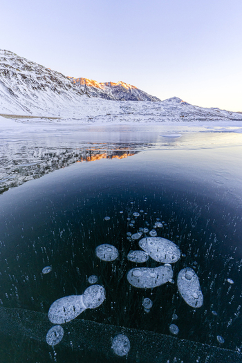 View of frozen lake with methane bubbles trapped in ice and snow covered mountains at sunset Madesimo Lombardy Italy. View of frozen lake with methane bubbles trapped in ice and snow covered mountains at sunset Madesimo Lombardy Italy.