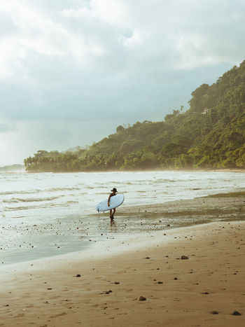 View of a surfer carrying a surfboard along a sandy beach with lush green hills and ocean waves in the background in Costa Rica.