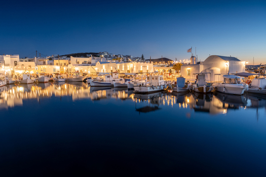 View of fishing boats moored in the harbor at dusk with illuminated white buildings reflecting in the calm water in Lefkada Greece.
