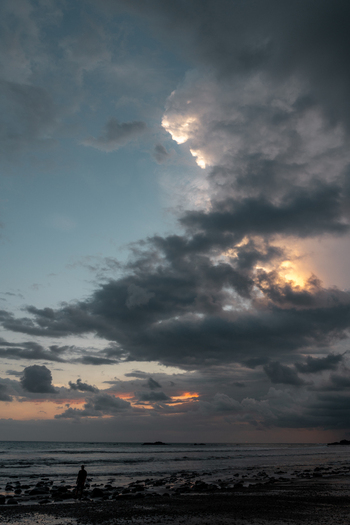 View of a lone person standing on a rocky beach at dusk with dramatic dark clouds and golden light breaking through over the ocean in Costa Rica.
