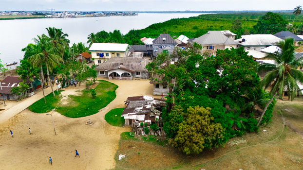 Aerial view of a riverside village with houses lush trees and people walking on a sandy path along the river in Minima Rivers Nigeria.