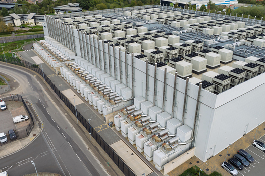 Aerial view of a large data center with numerous cooling units on the roof surrounded by perimeter fencing and parking areas in the United Kingdom.