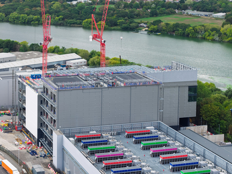 Aerial view of a large data center under construction with red cranes and colorful cooling units on the roof near a river in the United Kingdom.
