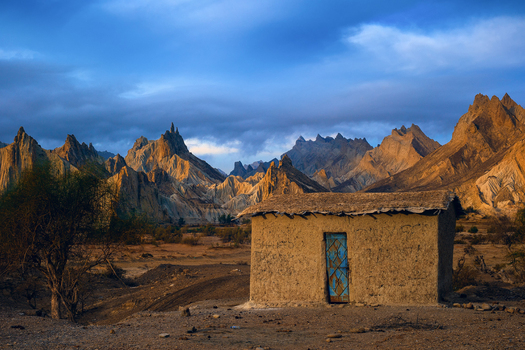 View of a small mud brick house with a blue door set against the dramatic jagged peaks of the Hingol mountains in a desert landscape in Awaran Balochistan Pakistan.