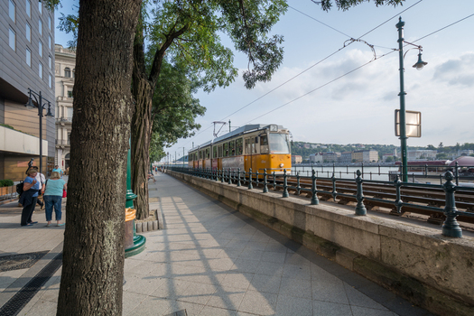 View of a yellow tram moving along the tracks on the Danube riverbank with pedestrians walking nearby in Budapest Hungary.