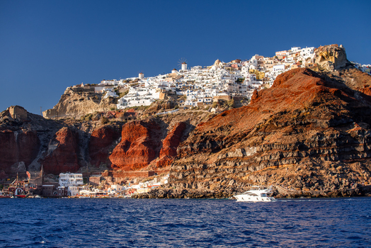 View of the white buildings of Fira perched on the volcanic cliffs of Santorini above the Aegean Sea with a boat in the foreground in Fira Greece. View of the white buildings of Fira perched on the volcanic cliffs of Santorini above the Aegean Sea with a boat in the foreground in Fira Greece.
