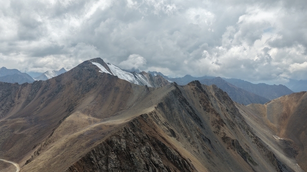 Aerial view of the rugged Enilchek mountain range with snow capped peaks and rocky slopes under a dramatic cloudy sky in Enilchek Issyk Kul Region Kyrgyzstan. Aerial view of the rugged Enilchek mountain range with snow capped peaks and rocky slopes under a dramatic cloudy sky in Enilchek Issyk Kul Region Kyrgyzstan.