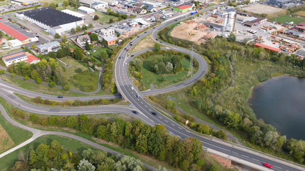 Aerial view of a highway interchange with a roundabout surrounded by lush green trees a small lake and industrial buildings in the background in Grossheubach Bavaria Germany. Aerial view of a highway interchange with a roundabout surrounded by lush green trees a small lake and industrial buildings in the background in Grossheubach Bavaria Germany.