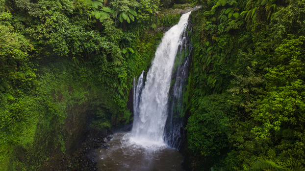 Aerial view of Gomblang Waterfall cascading down a steep cliff surrounded by dense tropical rainforest and lush green foliage in Kecamatan Kedungbanteng Jawa Tengah Indonesia. Aerial view of Gomblang Waterfall cascading down a steep cliff surrounded by dense tropical rainforest and lush green foliage in Kecamatan Kedungbanteng Jawa Tengah Indonesia.