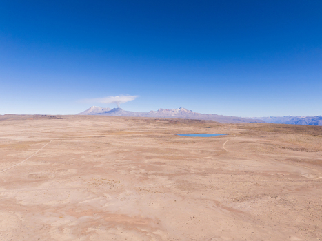 Aerial view of Mirador de los Andes featuring a vast arid landscape with a small blue lake and distant smoking volcanic peaks under a clear blue sky in Caylloma Arequipa Peru.