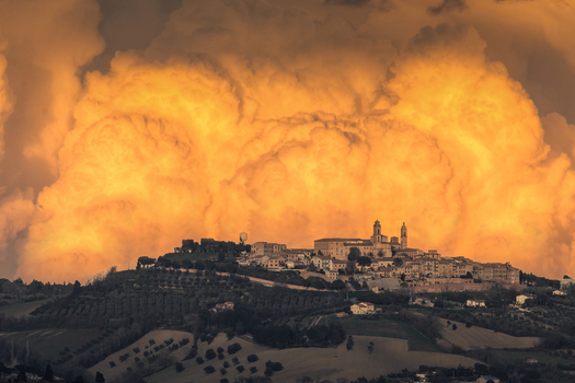 View of the historic hilltop town of Montecosaro illuminated by the warm golden light of a dramatic sunset with massive storm clouds looming in the background Montecosaro Marche Italy. View of the historic hilltop town of Montecosaro illuminated by the warm golden light of a dramatic sunset with massive storm clouds looming in the background Montecosaro Marche Italy.